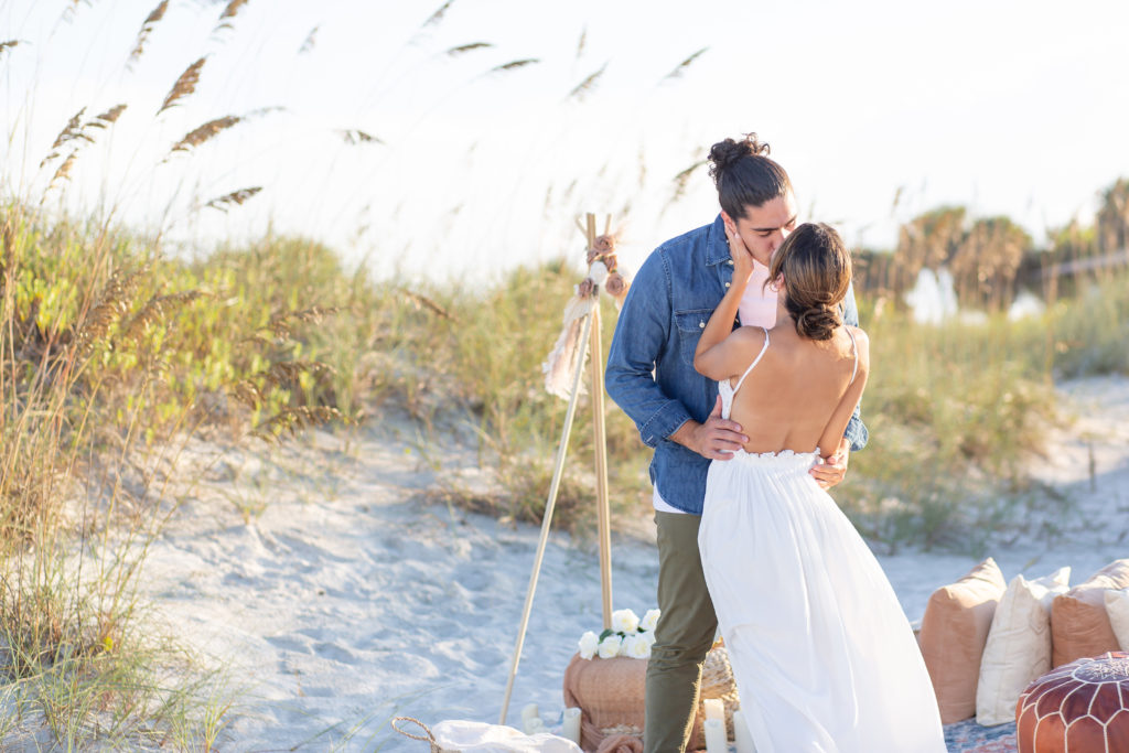 Newly engaged couple kisses in front of their picnic proposal setup on the beach