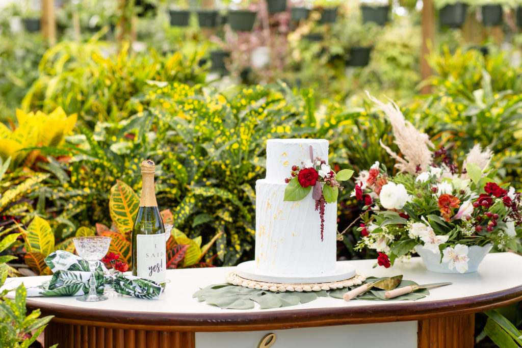 Greenhouse wedding tablescape with a white cake, champagne and a red-toned bouquet