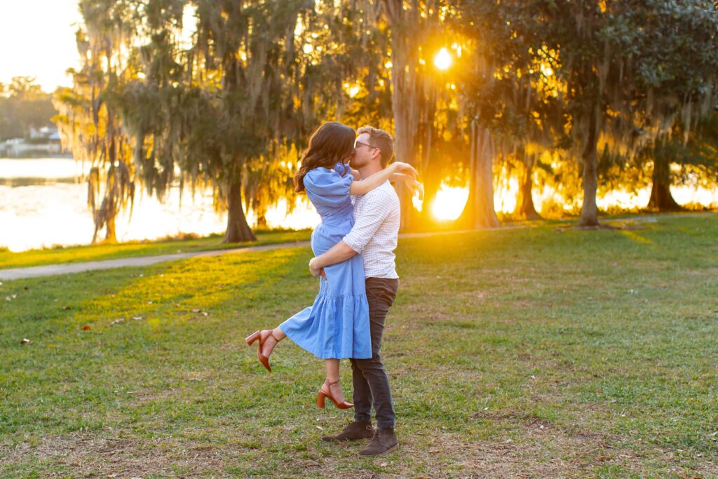 Guy lifts girl in front of lake and trees with the sunset peaking through for their engagement photos at Cypress Grove Park in Orlando, Florida