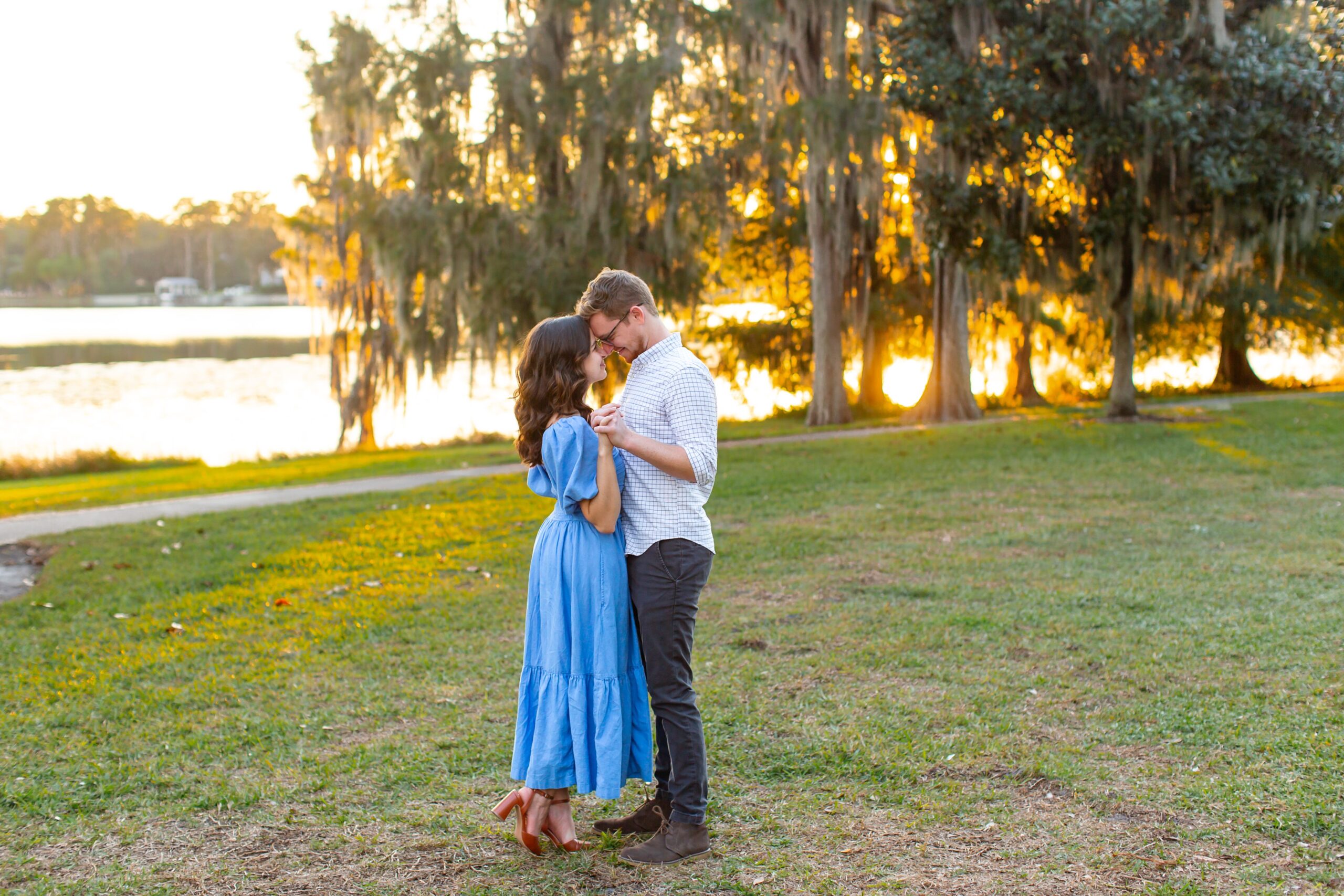 Couple stands and holds hands in front of lake and trees with the sunset peaking through for their engagement photos at Cypress Grove Park in Orlando, Florida