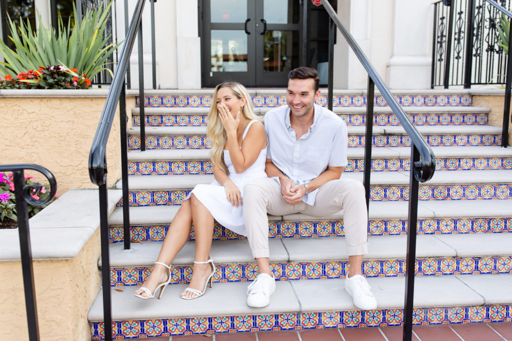 A woman in a white knee-length dress laughs while sitting next to her fiance on tiled stairs