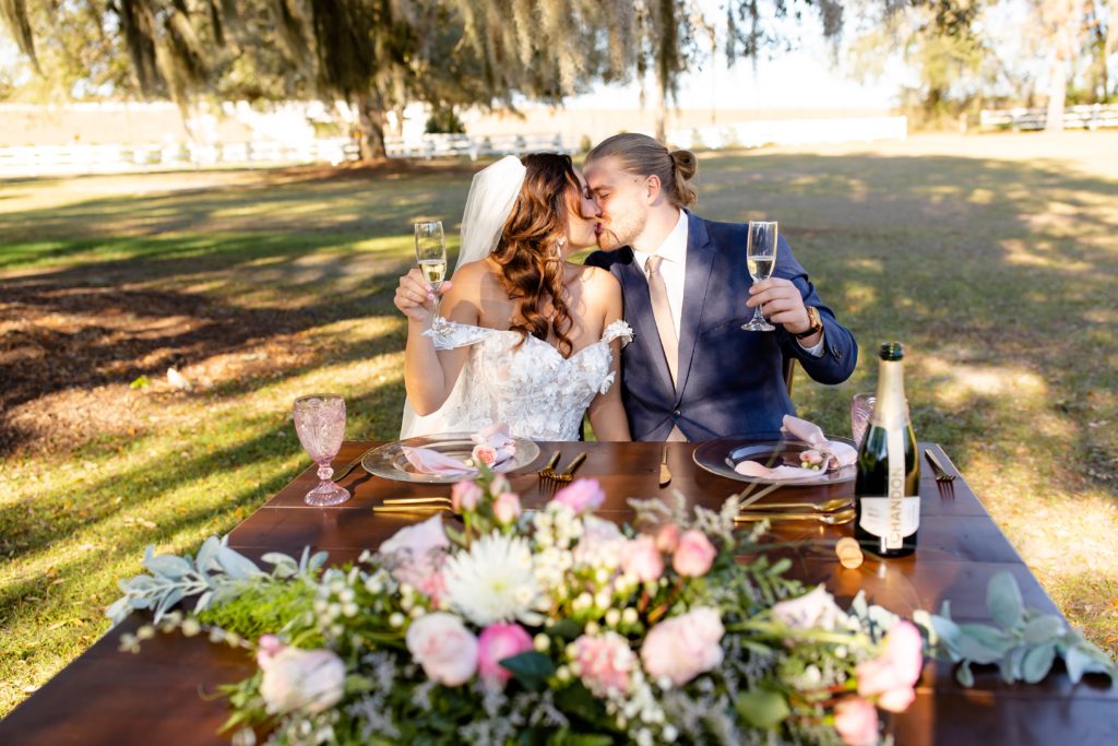 Bramble Tree Estate Wedding Photo — Bride and Groom kissing at sweet heart table with champagne under Spanish Moss tree