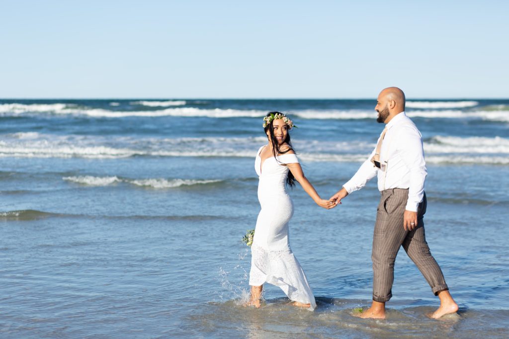 New Smyrna Beach Elopement Photo — Bride with flower crown holding hands with Groom and walking in the water on the beach