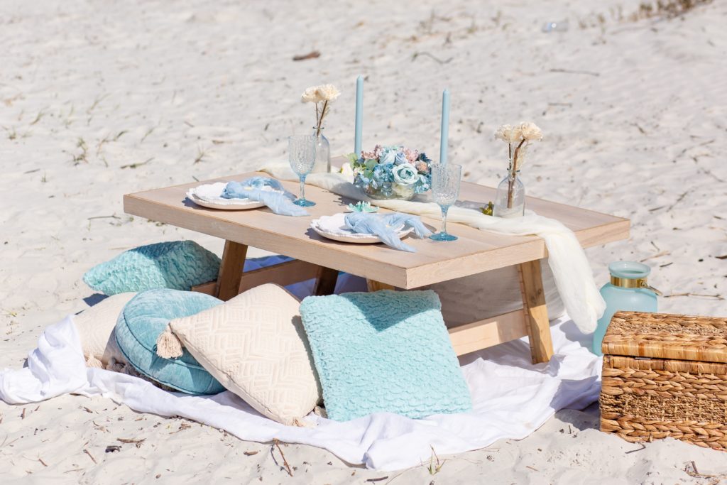 Elopement picnic on the beach with pastel blue pillows, a white blanket and a bleached wood table