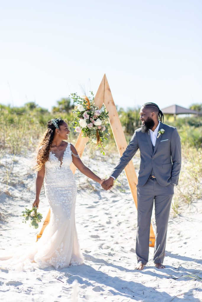 New Smyrna Beach Wedding Photo — Bride & Groom standing under arch holding hands and smiling at each other on the beach