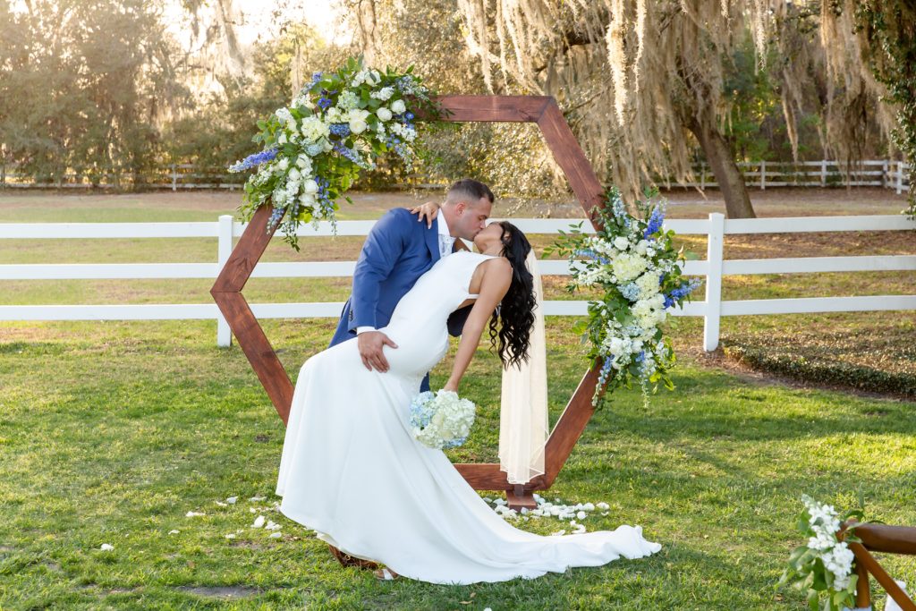 Bramble Tree Estate Wedding in Orlando, FL — 90 Day Fiance Wedding - Groom dipping and kissing bride at sunset under floral hexagon arch and hanging moss