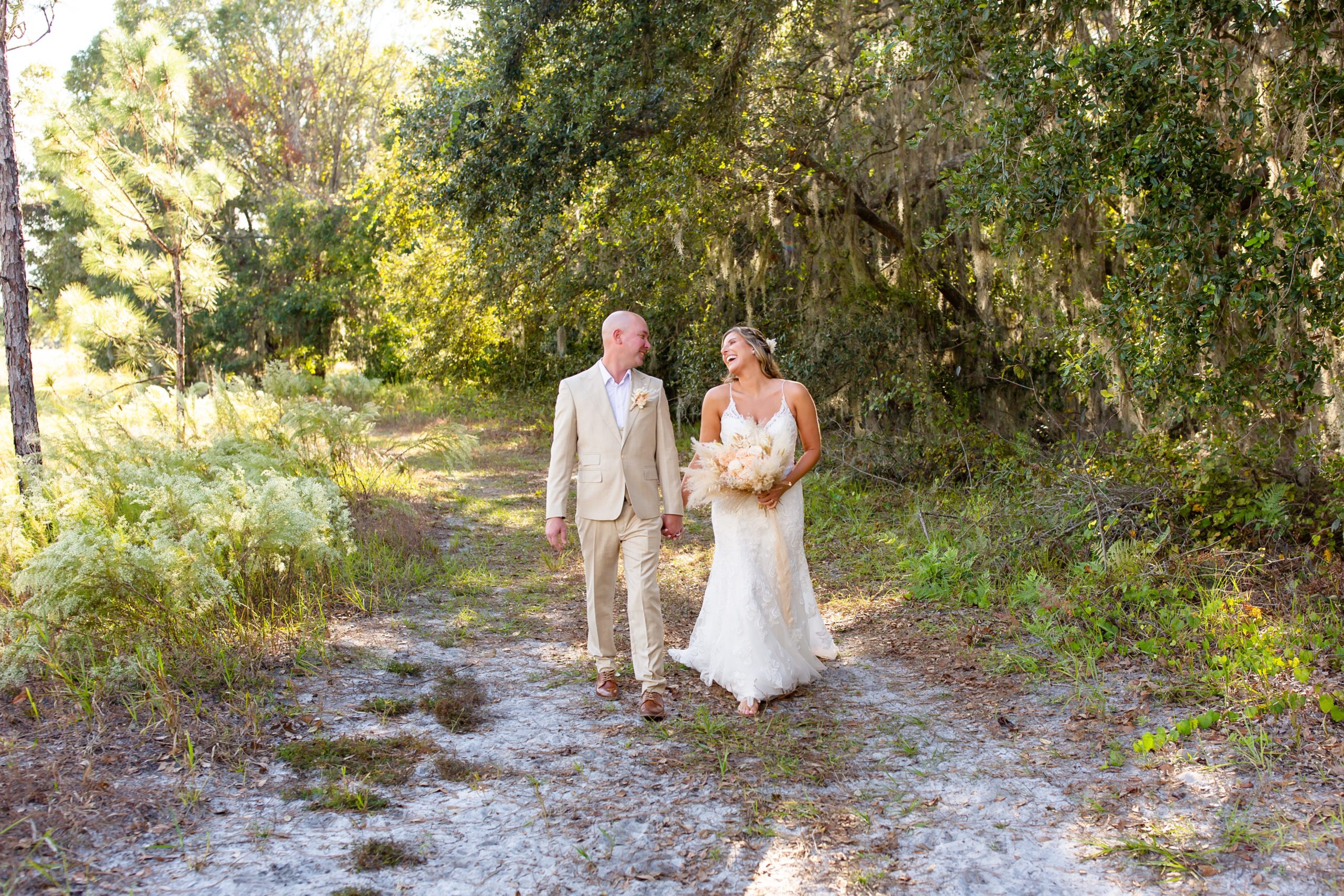 Lake Louisa Wedding Photos in Orlando, FL — Groom and Bride holding a dried floral bridal bouquet walking on path in field with hanging moss