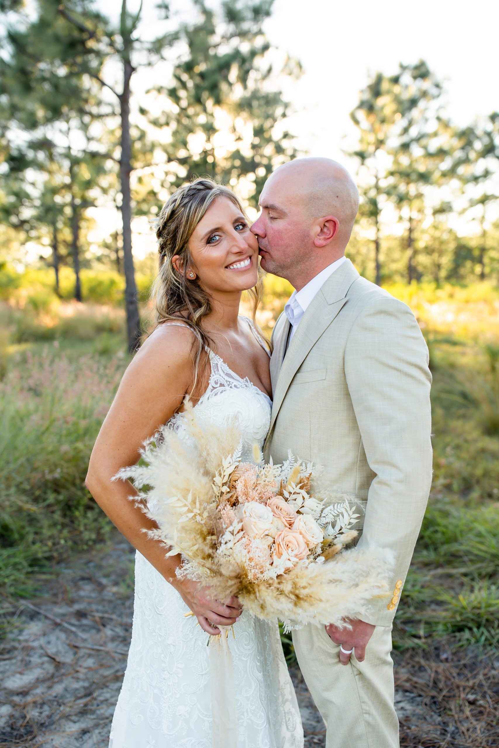 Lake Louisa Wedding Photos in Orlando, FL — Groom kissing Bride's cheek holding a dried bridal bouquet standing in field