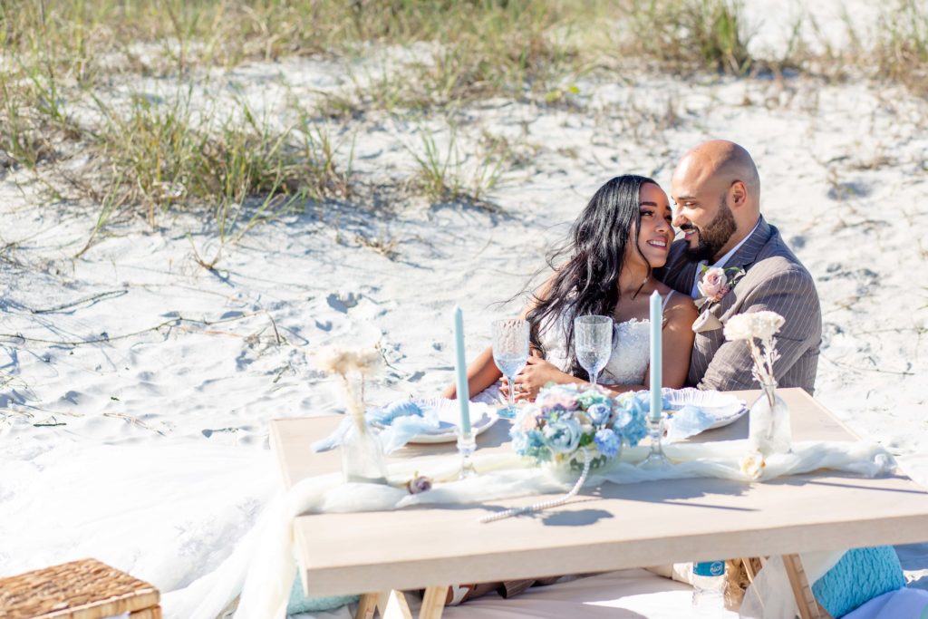 Bride and Groom sit on beach with small picnic table with pastel table settings in New Smyrna Beach, Florida