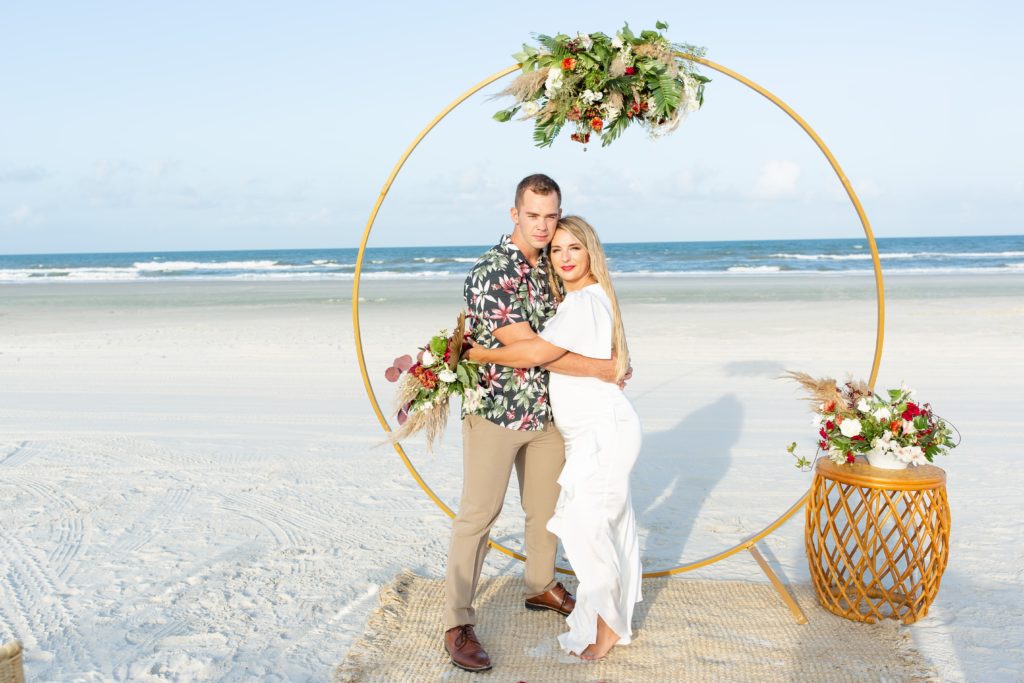 Bride & Groom standing under metal circle ceremony arch with white florals at the top on the beach
