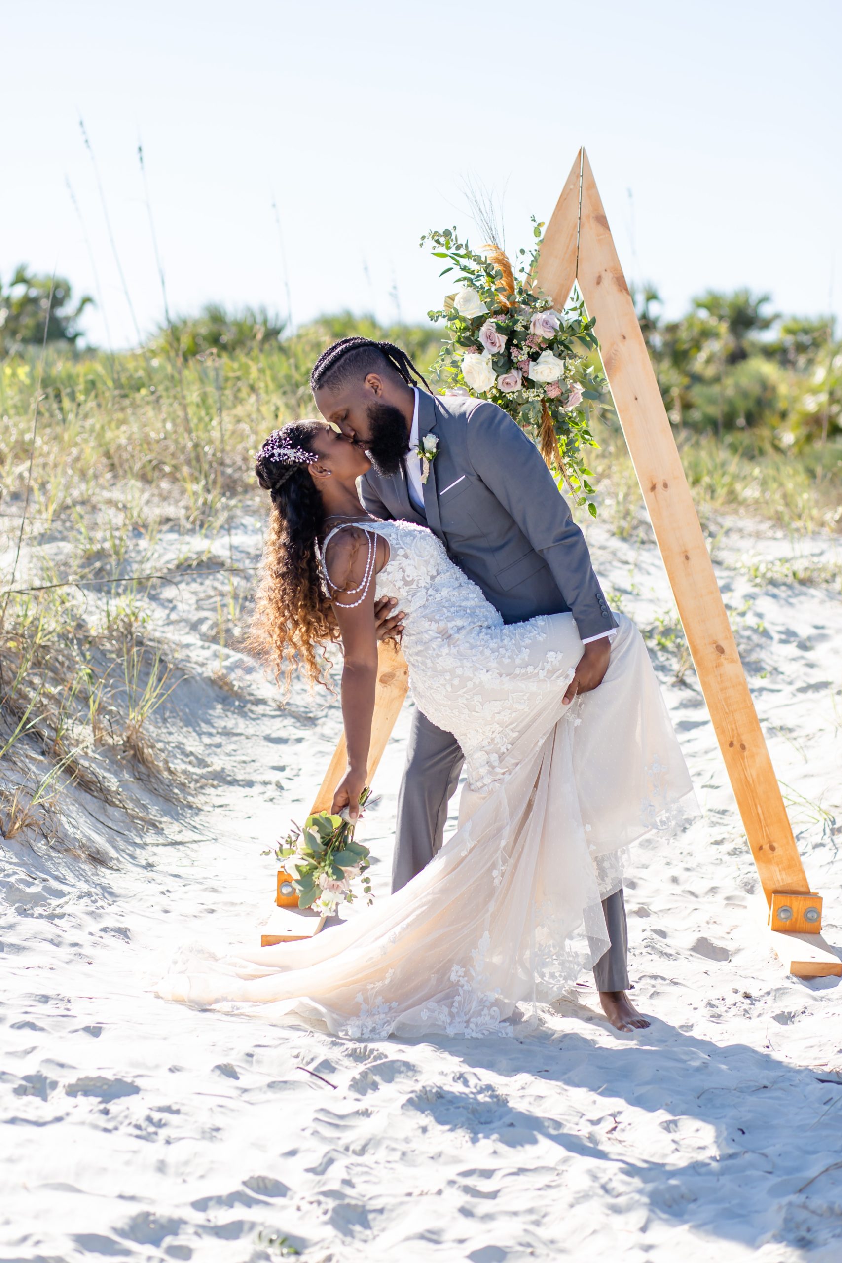Bride & Groom kissing under wooden ceremony triangle arch with floral accents on the beach