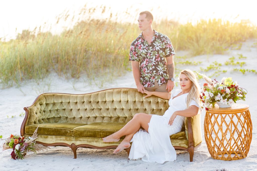 Bride sitting on green couch on the beach with side table of flowers while groom stands behind her on the beach in St. Augustine, FL