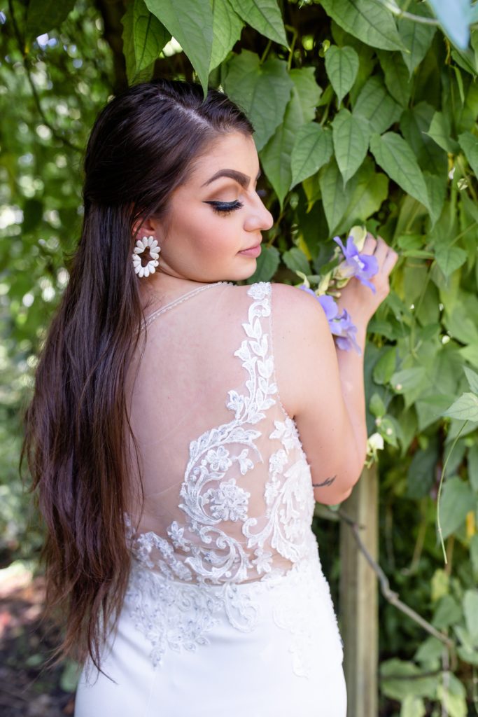 Bride wearing lace bridal gown and white clay earrings holding purple flowers by ivy wall at Mead Botanical Garden in Orlando, FL