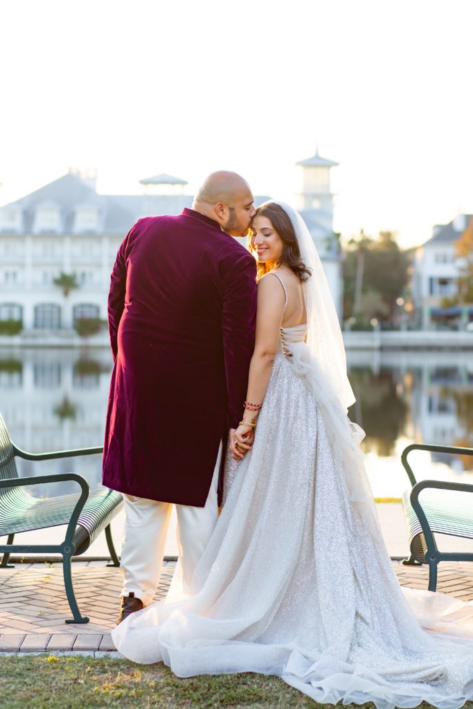 Groom in Purple suit kissing Bride's Forehead wearing a sequin bridal gown with veil standing by lake in front of Celebration Florida Bohemian Hotel