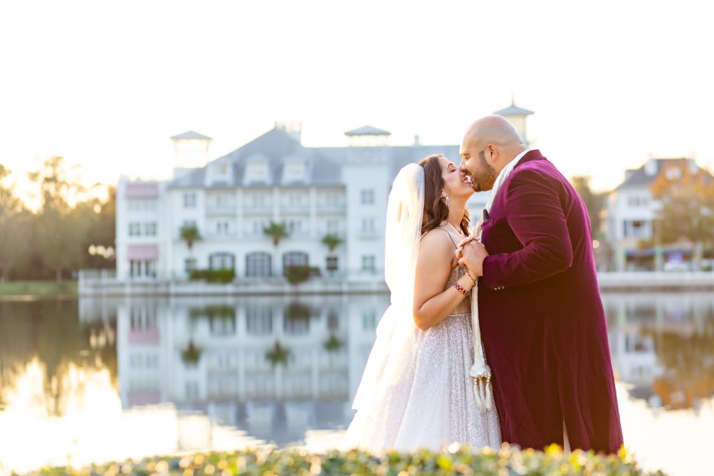 Groom in Purple suit kissing Bride wearing a sequin bridal gown with veil standing by lake at sunset in front of Celebration Florida Bohemian Hotel