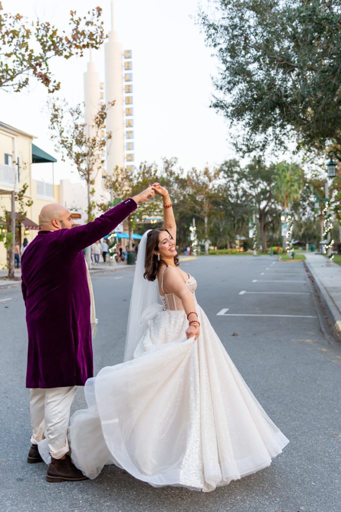 Groom in Purple suit twirling Bride wearing a sequin bridal gown with veil standing in street of Celebration Florida by cinema