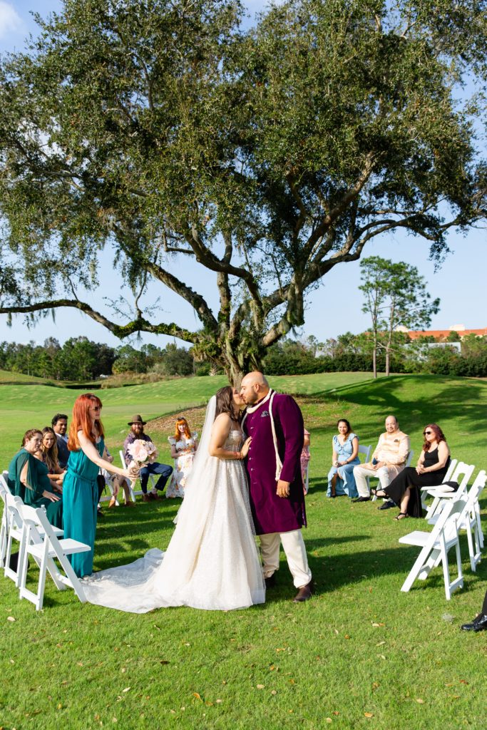 Bride & Groom first kiss at wedding ceremony under large tree with guests in circle around them at Celebration Golf Club