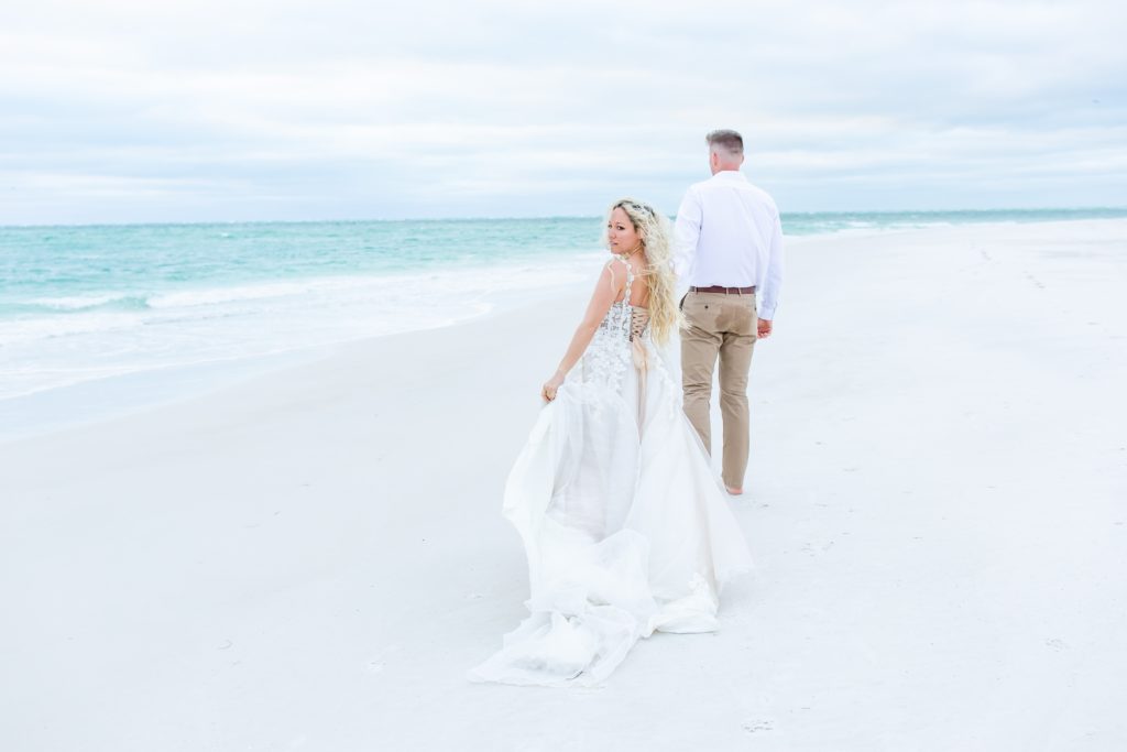 Groom walking with bride wearing cascading bridal gown on the beach on Anna Maria Island, FL