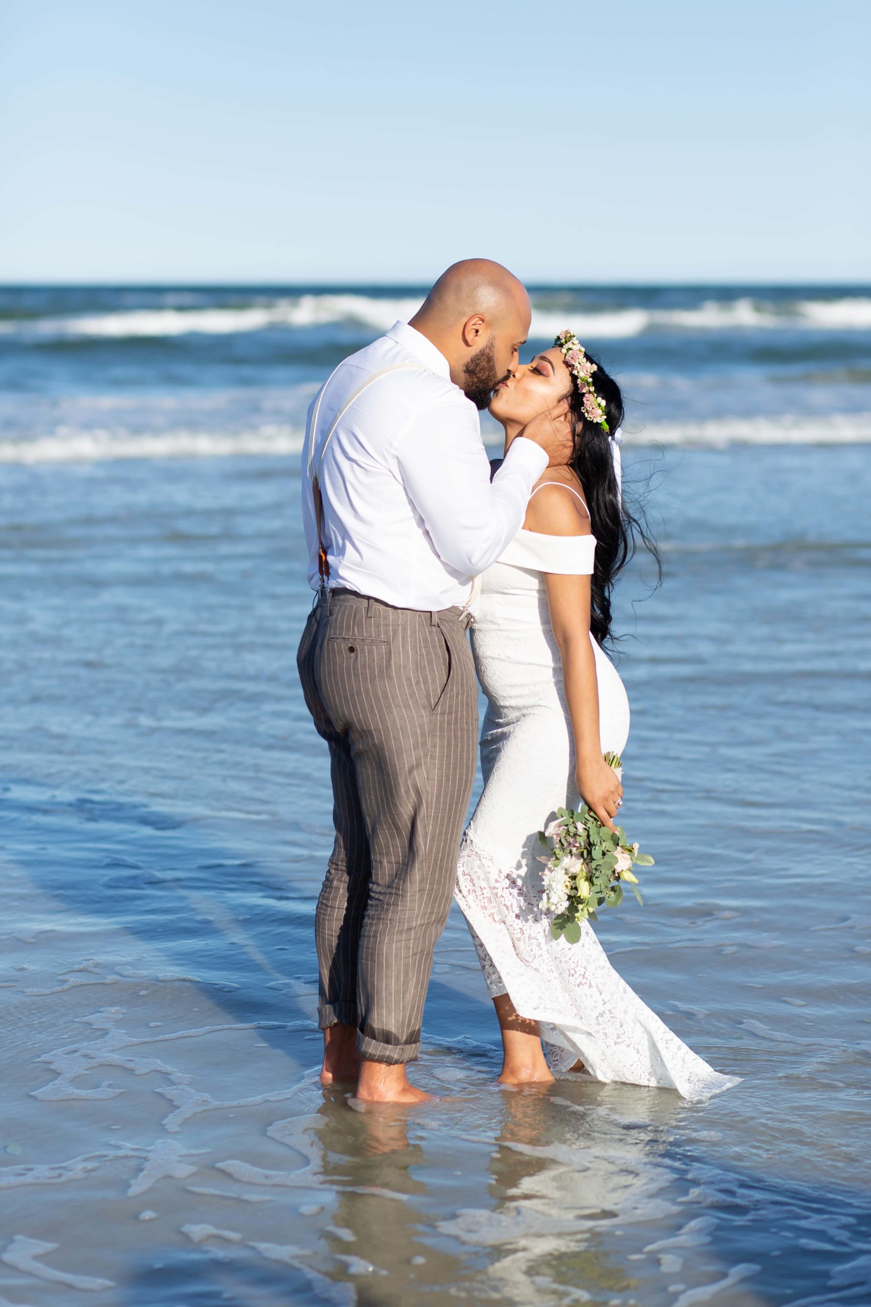 Groom kissing bride standing in ocean with bride wearing floral crown and petite bridal bouquet in New Smyrna Beach, FL
