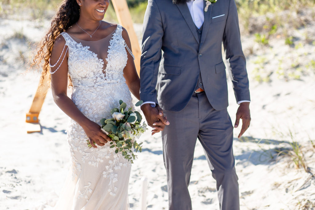 Bride holding petite bouquet holding hands with groom on beach in front of triangle arch at New Smyrna Beach, FL