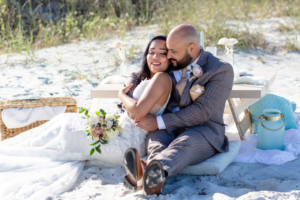 Bride and Groom hugging and sitting in front of pop up beach picnic while bride holds bouquet at New Smyrna Beach Elopement