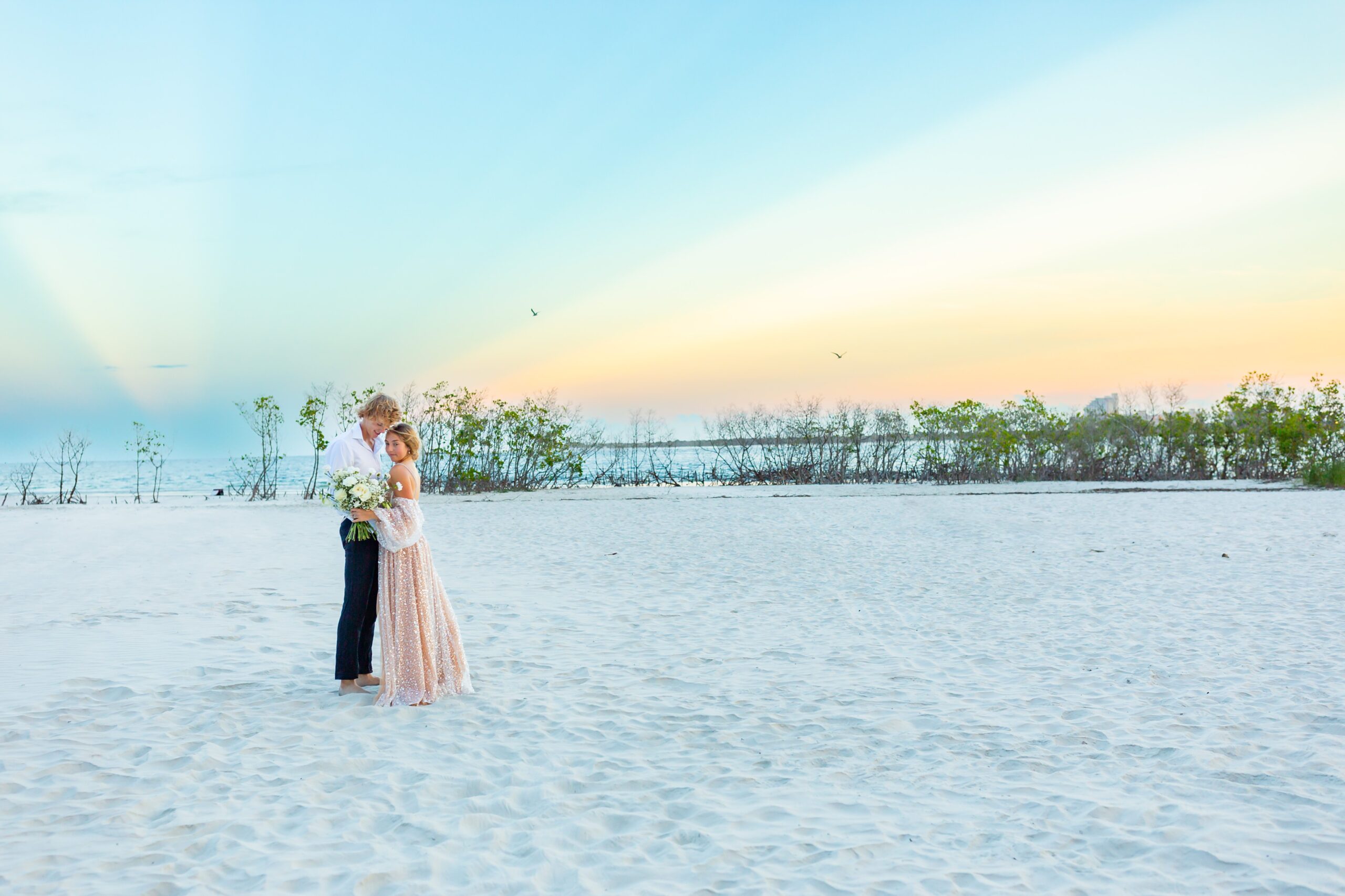 Bride and Groom stand on white sand beach with stunning colors of sunset rays behind them at Ponce Inlet Beach