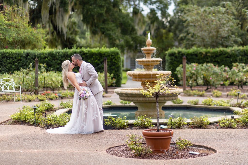 Groom dipping and kissing bride standing in front of fountain in rose garden at Leu Gardens in Orlando, FL