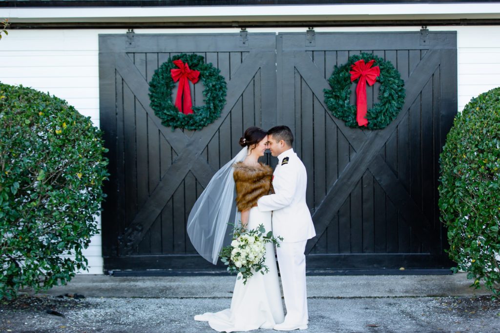 Bride wearing fur shawl & Groom laughing in front of black barn door with Christmas Wreaths at Bramble Tree Estate in Orlando, Florida