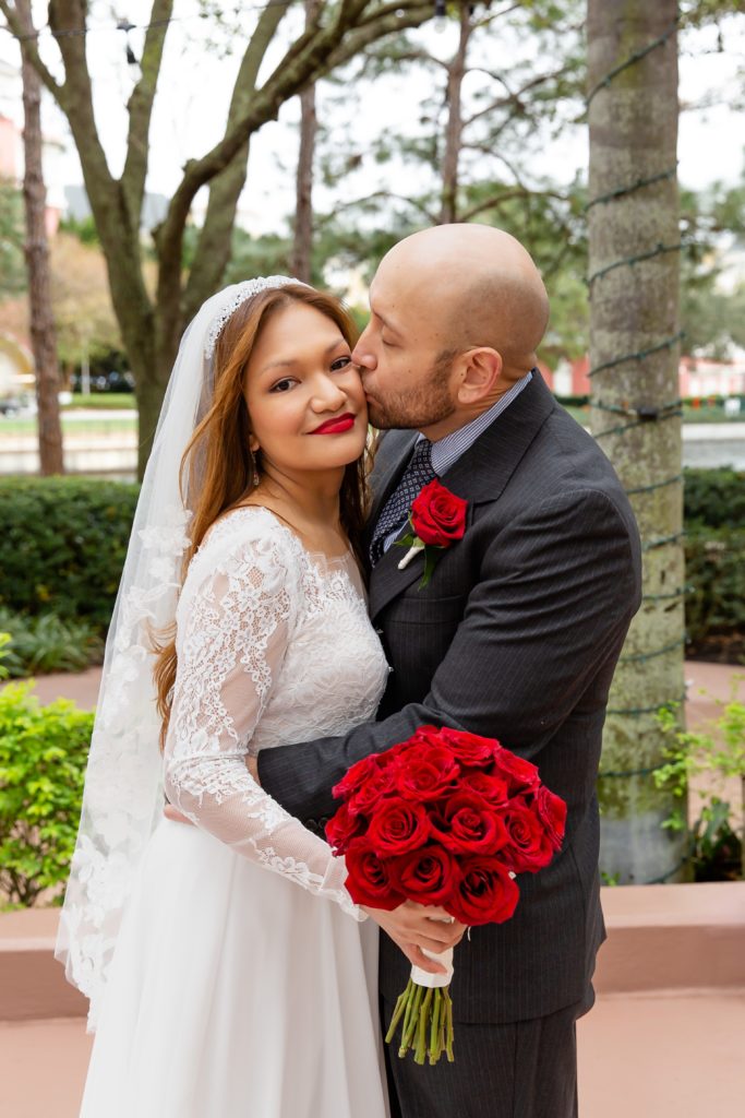 Groom kissing Bride's cheek with red rose bouquet after eloping at Disney Swan Resort in Orlando, FL