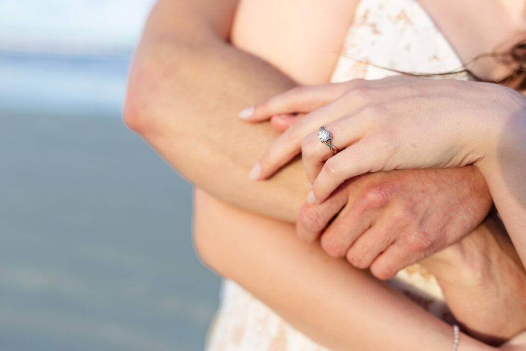 New Smyrna Beach Engagement Photo — Couple with arms wrapped around each other showing off custom designed engagement ring on the beach at sunset