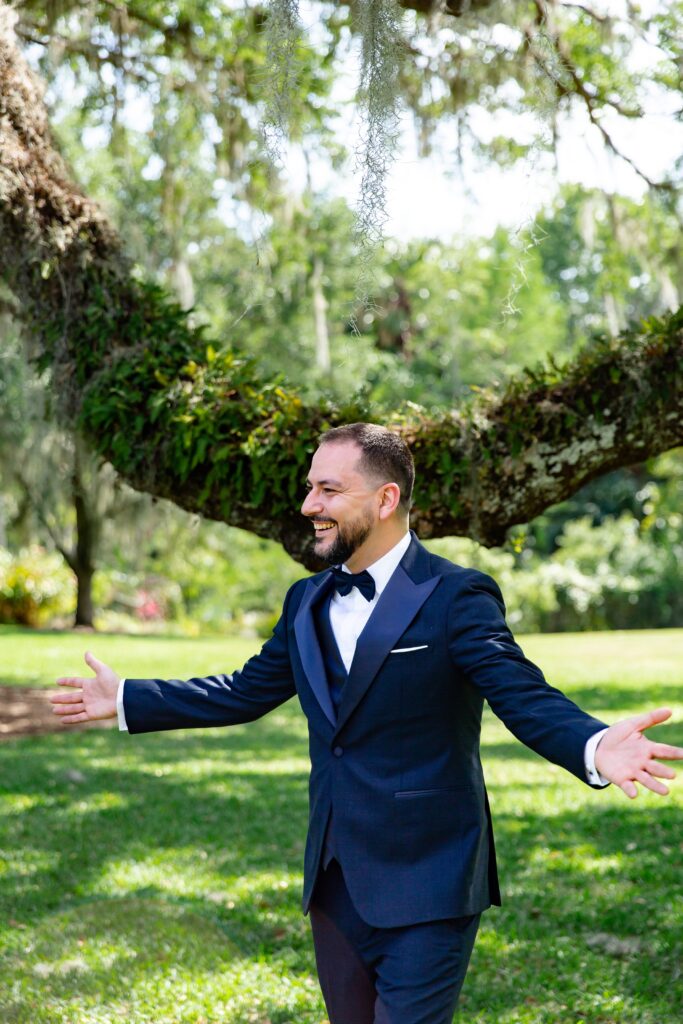 Groom in navy suit smiling when seeing bride at the first look under live oak tree at Leu Gardens Elopement in Orlando