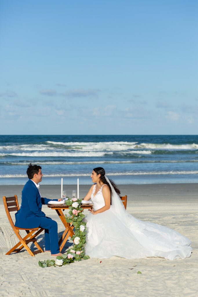 Bride and Groom sitting at wooden table for two on the beach with floral accents on the beach at their Florida Beach Wedding Packages