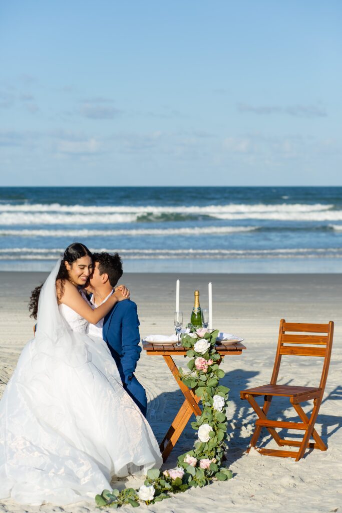 Bride sitting on Groom's lap at wooden table for two on the beach with floral accents on the beach included in their Florida Beach Wedding Packages