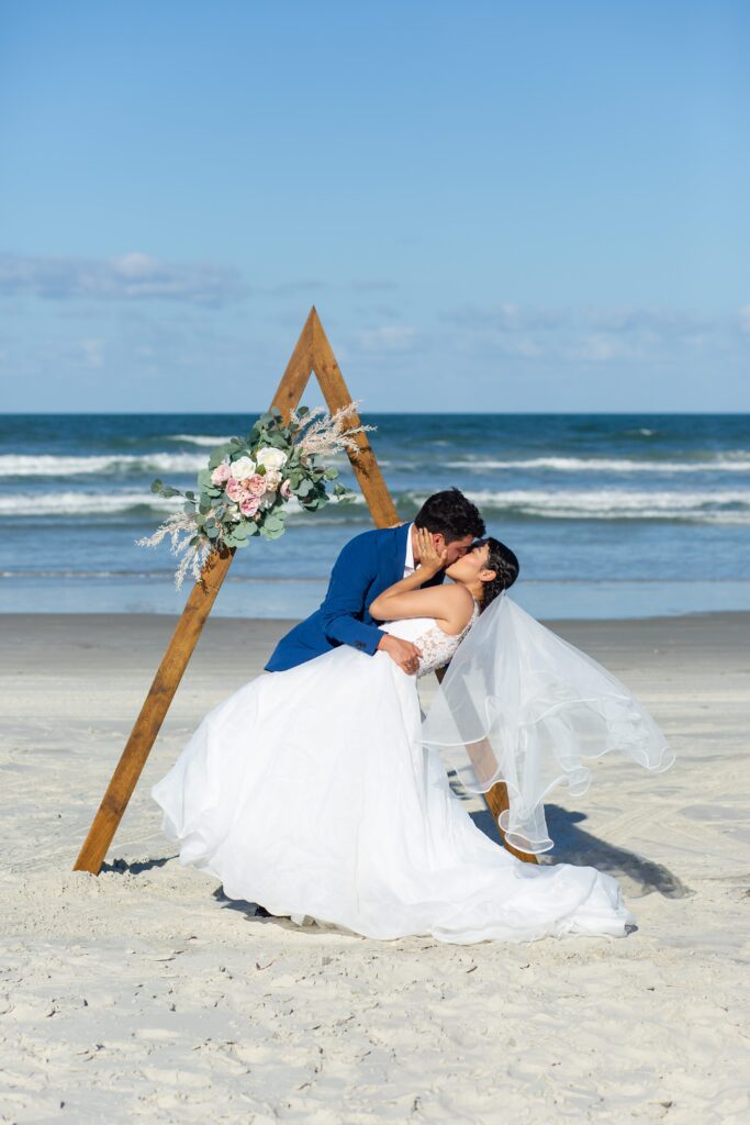 Groom dipping bride and kissing her under wooden ceremony triangle arch with floral accents on the beach with Florida Beach Wedding Packages