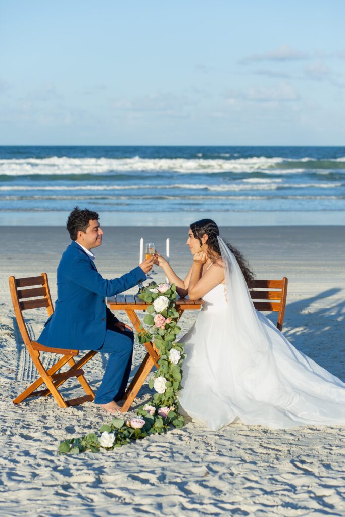 Bride and Groom sitting at wooden table for two on the beach toasting with floral accents on the beach included in their Florida Beach Wedding Packages