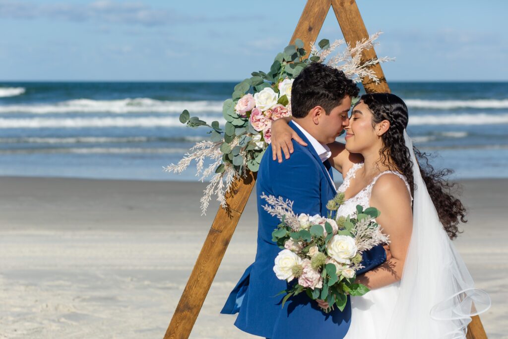 Bride and Groom kissing her under wooden ceremony triangle arch with floral accents on the beach included in their Florida Beach Wedding Packages