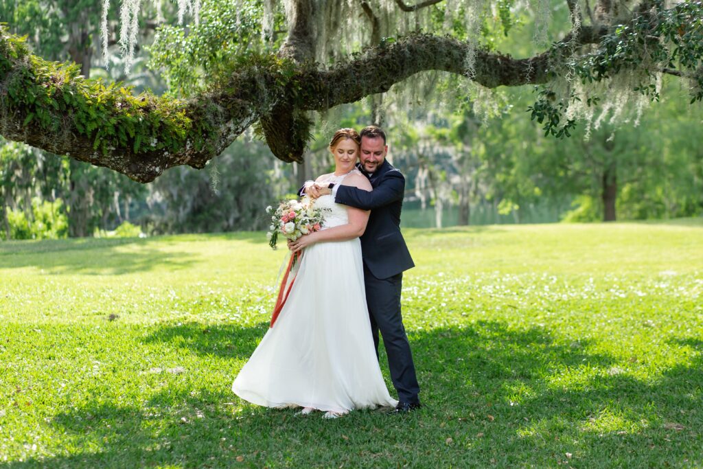 Groom in navy suit wraps arms around Bride holding bouquet under live oak tree after their elopement at Leu Gardens in Orlando