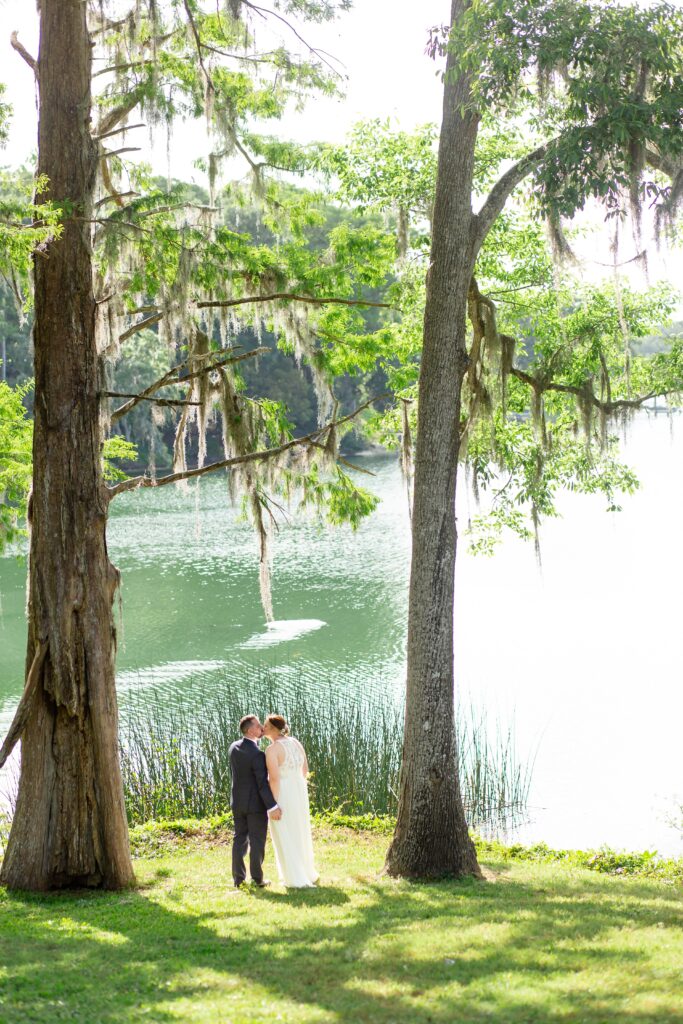 Bride and Groom kissing and standing next to lake under live oak tree after their elopement at Leu Gardens in Orlando