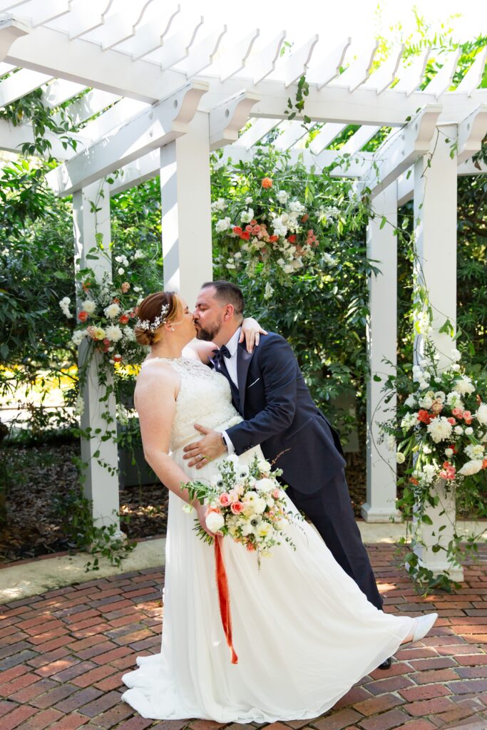 Groom in navy suit dipping Bride holding bouquet under trellis after their elopement at Leu Gardens in Orlando