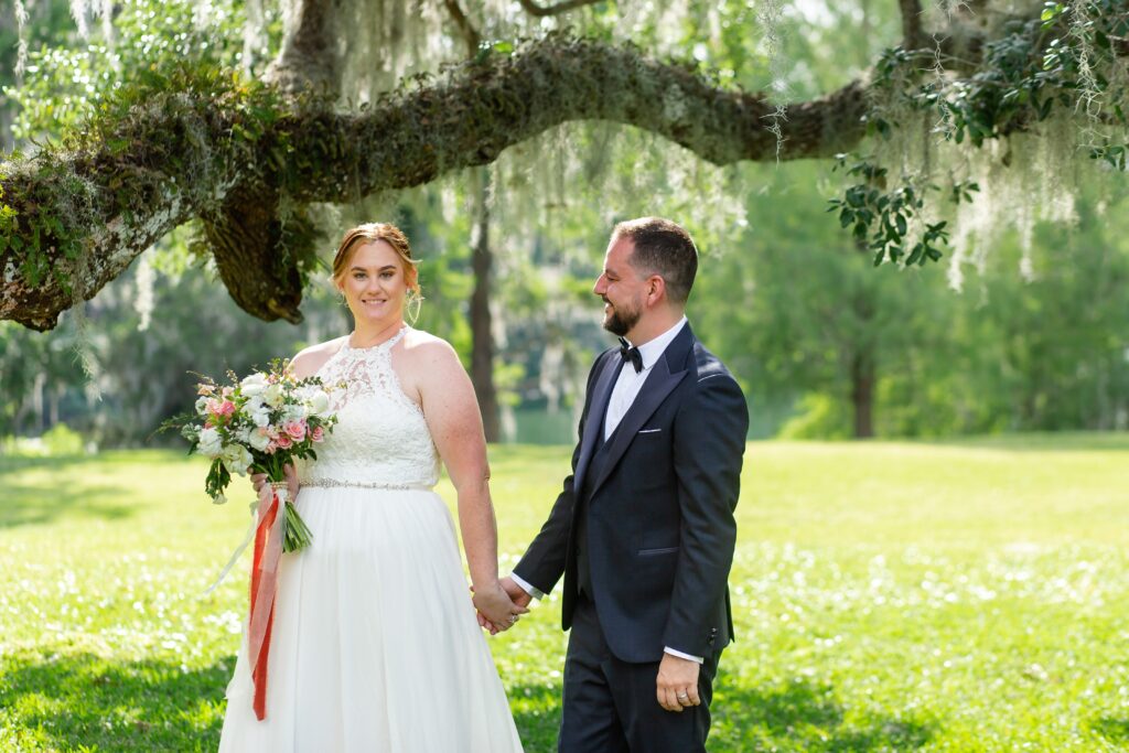 Groom in navy suit walks with Bride holding bouquet under live oak tree after their elopement at Leu Gardens in Orlando