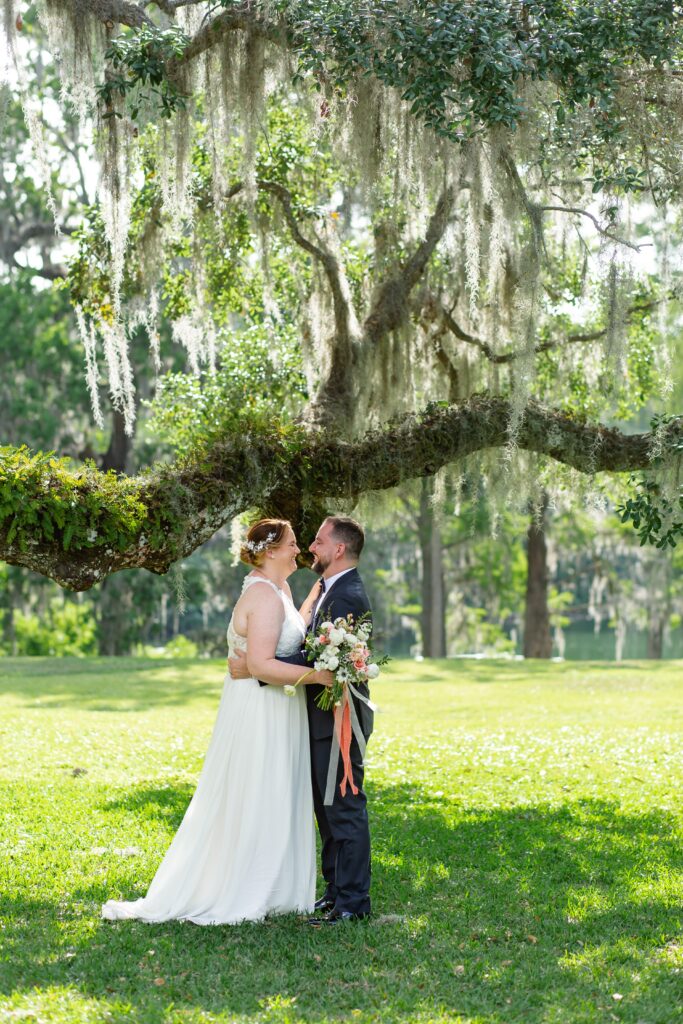 Groom in navy suit laughs with Bride holding bouquet under live oak tree after their elopement at Leu Gardens in Orlando