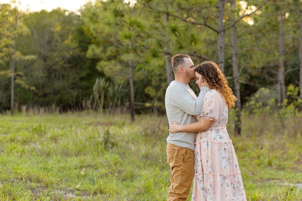 Guy in gray shirt kisses forehead and wraps arms around girl wearing light pink maxi dress in field for their engagement photos at Lake Louisa State Park in Orlando