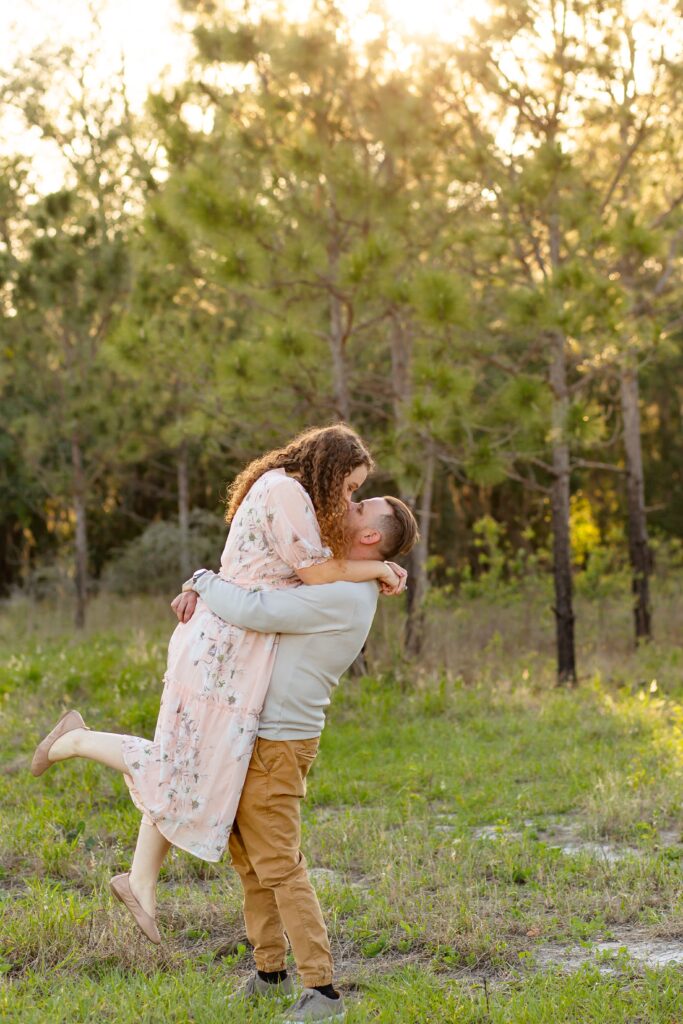 Guy in gray shirt lifts and kisses girl wearing light pink maxi dress in field for their engagement photos at Lake Louisa State Park in Orlando