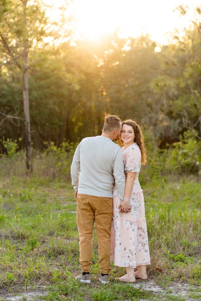 Guy in gray shirt holds hands and kisses girl wearing light pink maxi dress in field at sunset for their engagement photos at Lake Louisa State Park in Orlando