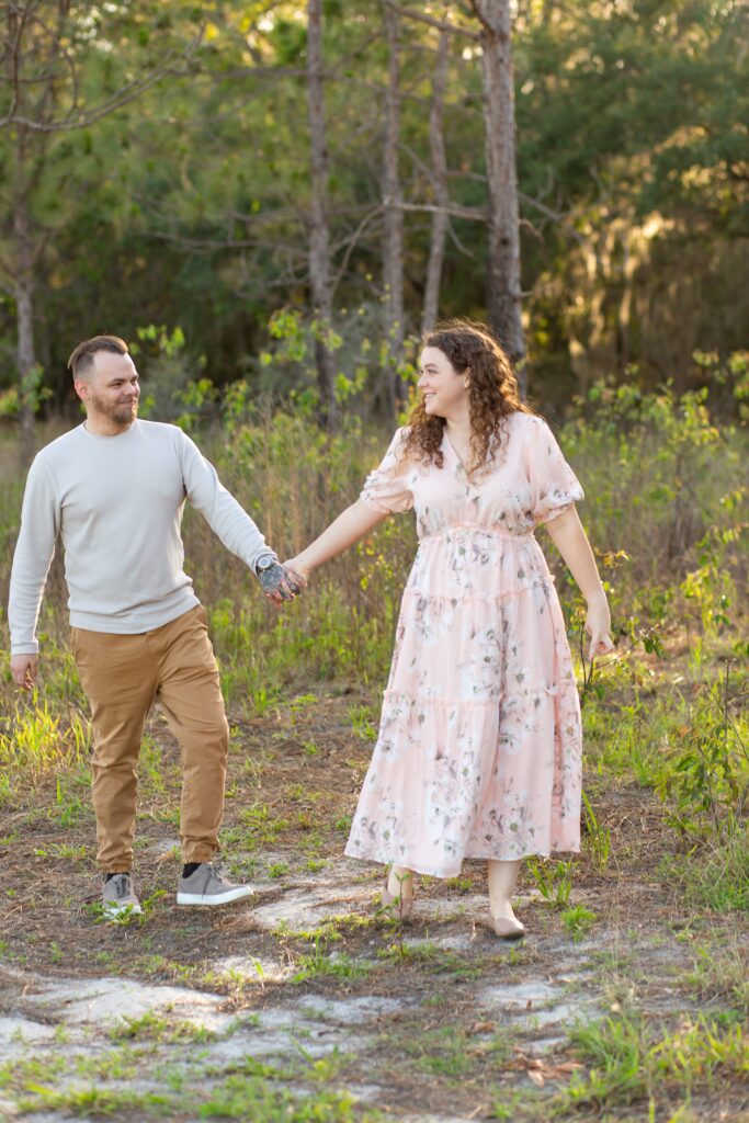 Girl wearing light pink maxi dress leads Guy in gray shirt walking in field at sunset for their engagement photos at Lake Louisa State Park in Orlando