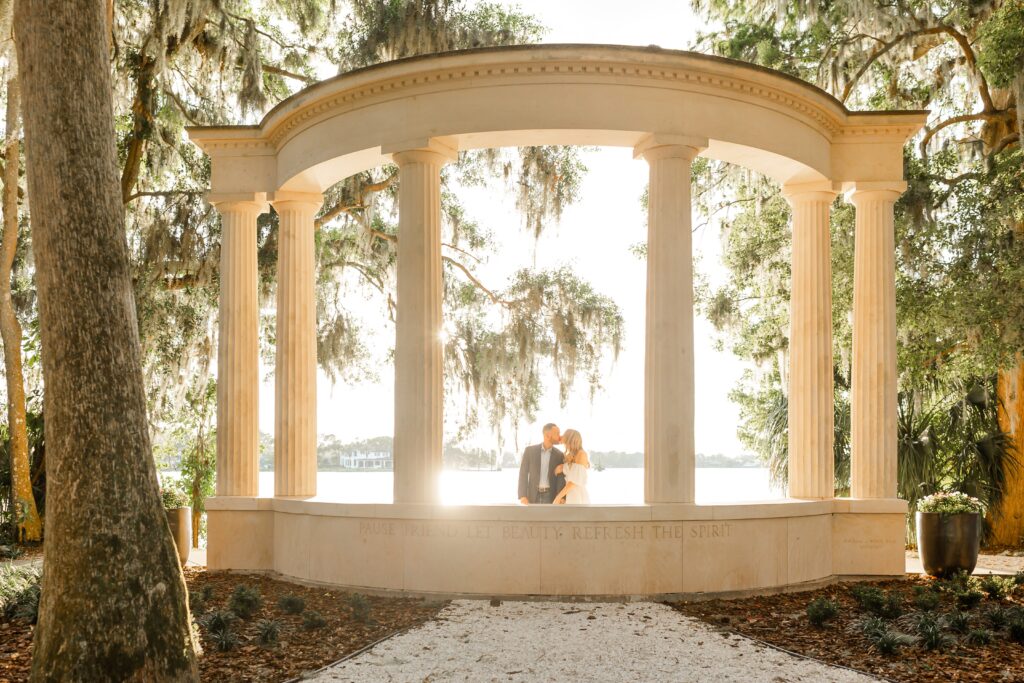 Couple kissing in front of columns in front of lake for their sunset Orlando Engagement Photos at Kraft Azalea Garden in Winter Park Florida