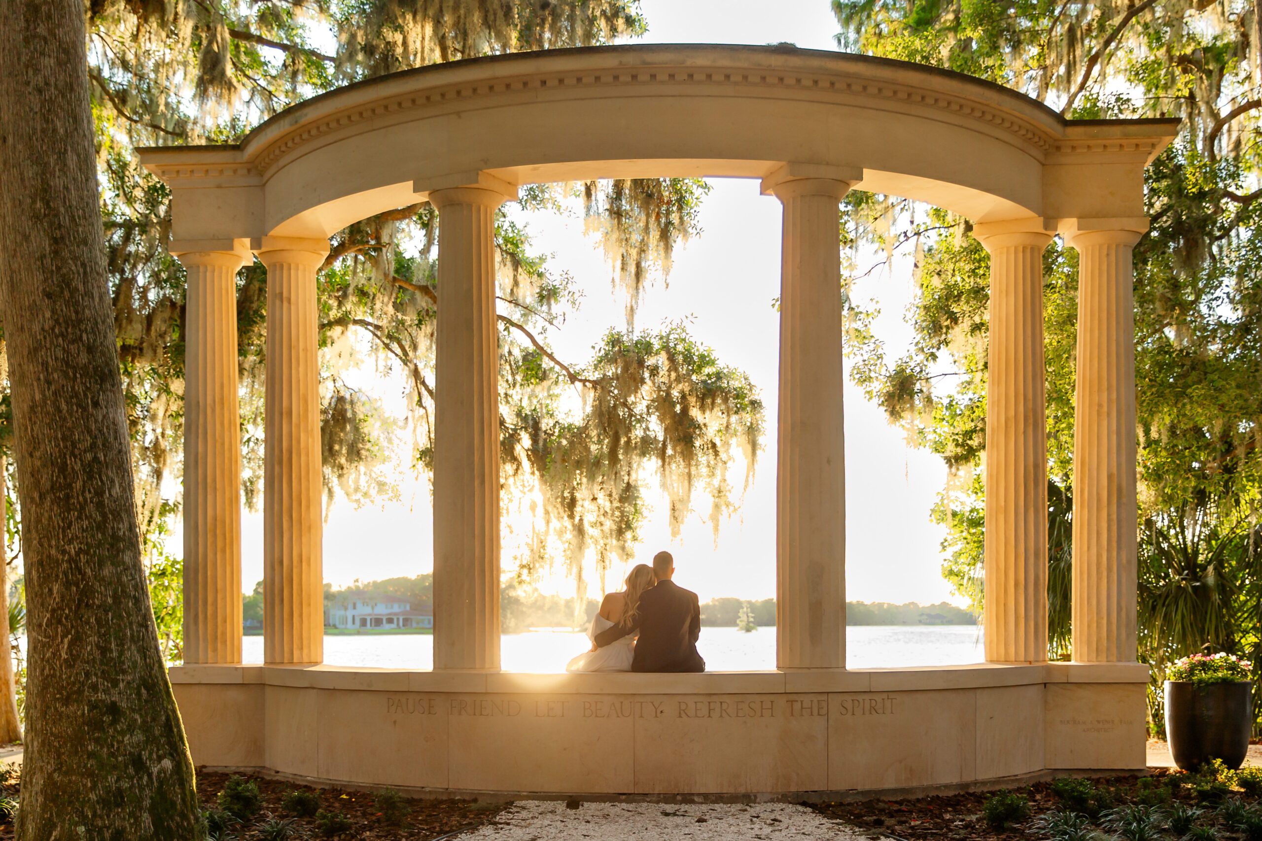 Guy snuggles girl while she lays her head on his shoulder sitting columns in front of lake for their sunset Orlando Engagement Photos at Kraft Azalea Garden in Winter Park Florida