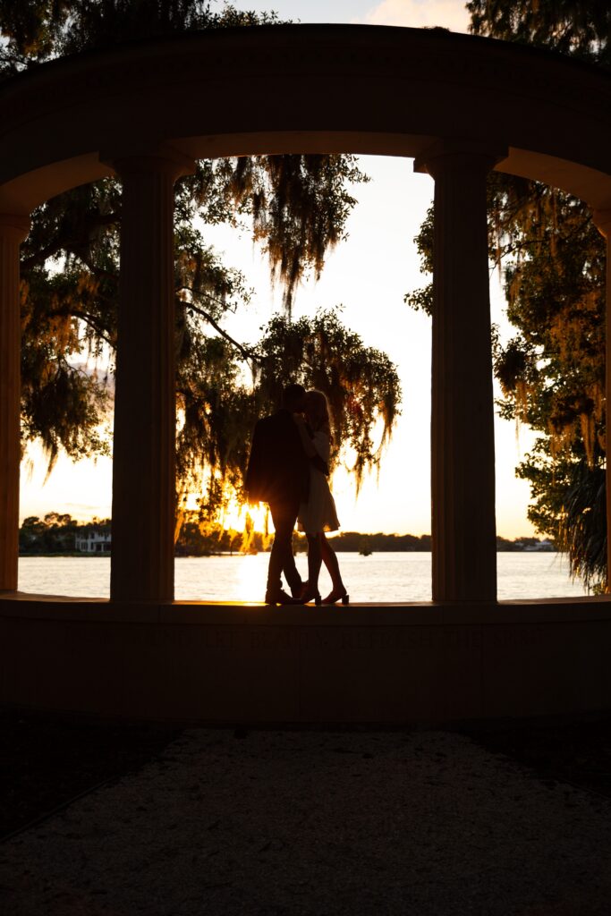 Silhouette of couple as they dance on columns in front of lake for their sunset Orlando Engagement Photos at Kraft Azalea Garden in Winter Park Florida