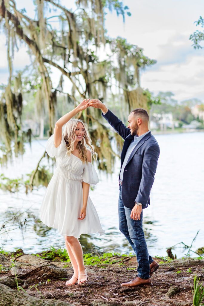 Guy twirls girl in front of lake with live oak tree for their sunset Orlando Engagement Photos at Kraft Azalea Garden in Winter Park Florida