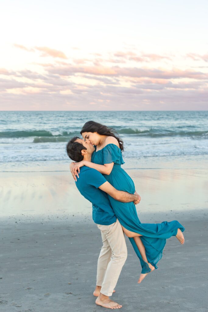 Guy lifts girl and kisses her at sunset on the beach for their engagement photos in Cocoa Beach Florida